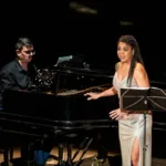 Chiara Santoro cantando em recital "As Brasileiras", partitura em sua frente e ela voltada ao público. Atrás dela está o pianista Silas Barbosa.