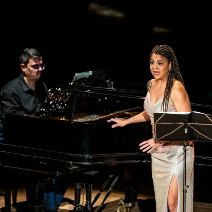 Chiara Santoro cantando em recital "As Brasileiras", partitura em sua frente e ela voltada ao público. Atrás dela está o pianista Silas Barbosa.