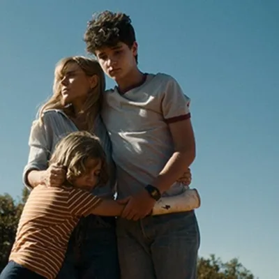 Pequenas Criaturas | Conheça o Filme Vencedor do Troféu Redentor de Melhor Longa de Ficção da Première Brasil Helena (Carolina Dieckmann) e seus filhos, André (Théo Medon) e Dudu (Lorenzo Mello) no filme "Pequenas Criaturas", todos abraçados em um céu azul com filtro que dá a impressão de um Brasil dos anos 80.