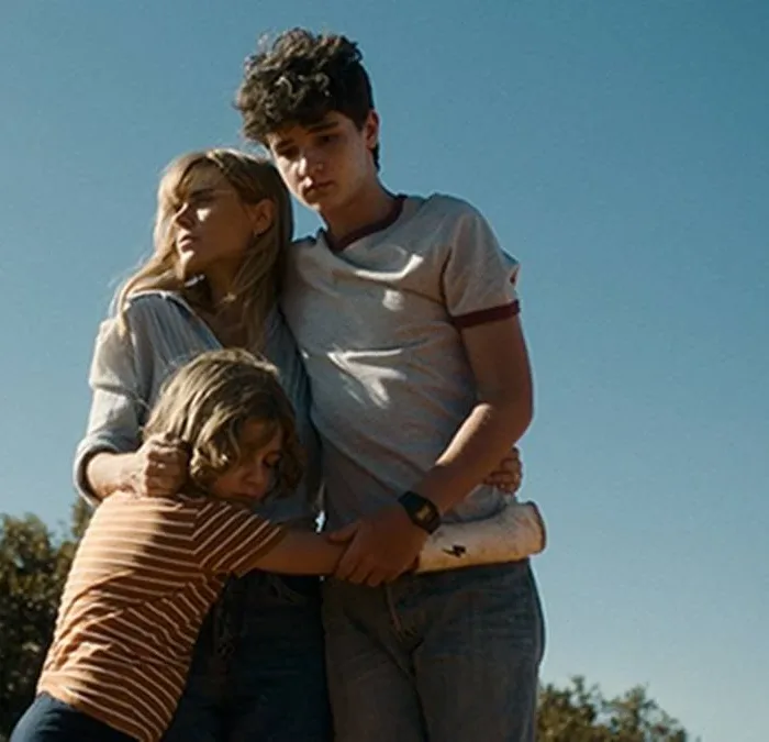 Helena (Carolina Dieckmann) e seus filhos, André (Théo Medon) e Dudu (Lorenzo Mello) no filme "Pequenas Criaturas", todos abraçados em um céu azul com filtro que dá a impressão de um Brasil dos anos 80.