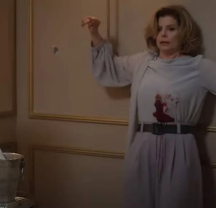 Cena da morte de Odete Roitman no remake de 2025 de "Vale Tudo", com Débora Bloch com o icônico vestido do original e deslizando na parede com a barriga ensanguentada.