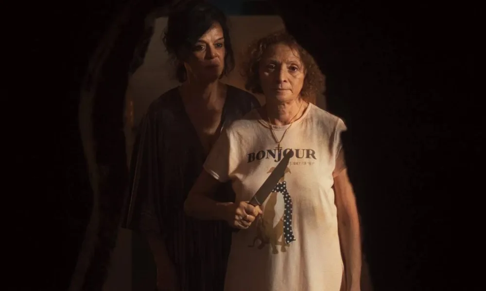 Imagem promocional do filme "Gravidade" (2025). Uma mulher à frente segura uma faca na altura do peito, enquanto outra está atrás dela, olhando para o mesmo ponto que ela, nosso ponto de vista de câmera.