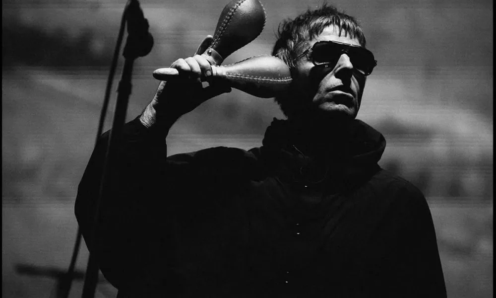 Liam Gallagher em show do Oasis na turnê de reunião de 2025, em Sidney, dia 7 de novembro, de jaqueta e óculos escuros, segurando duas maracas. Foto em preto e branco.