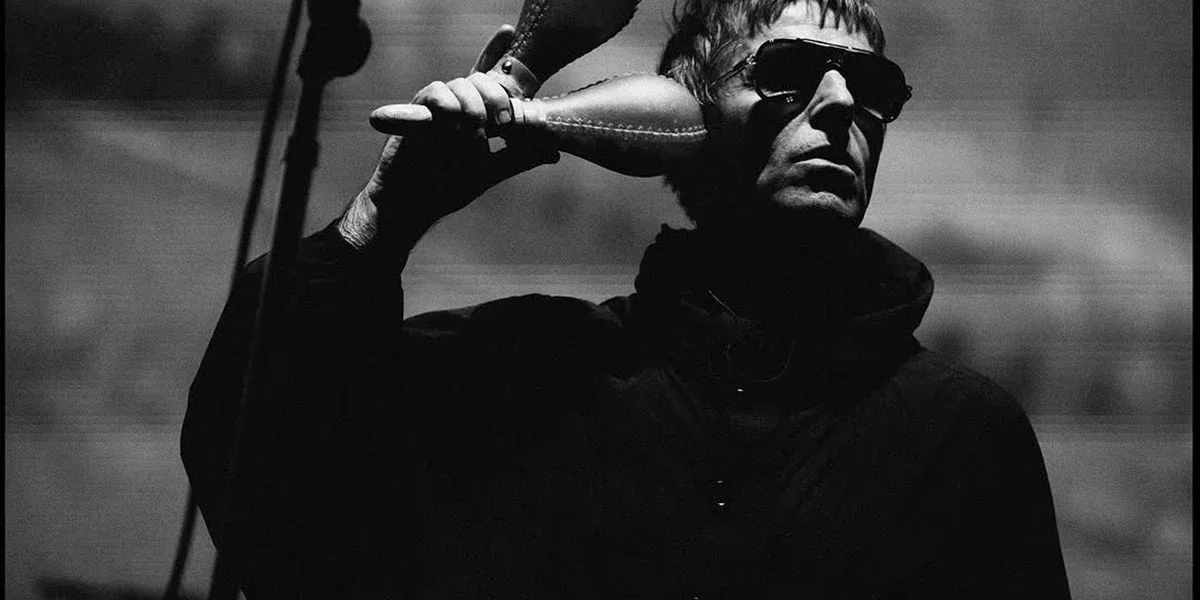 Liam Gallagher em show do Oasis na turnê de reunião de 2025, em Sidney, dia 7 de novembro, de jaqueta e óculos escuros, segurando duas maracas. Foto em preto e branco.