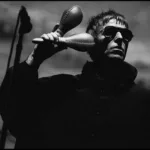 Liam Gallagher em show do Oasis na turnê de reunião de 2025, em Sidney, dia 7 de novembro, de jaqueta e óculos escuros, segurando duas maracas. Foto em preto e branco.