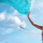 Catherine Laga‘aia como Moana no live action de 2026. Ela está no canto inferior direito tocando o mar que se move até ela como um tentáculo brilhante, ocupando a maior parte da tela.