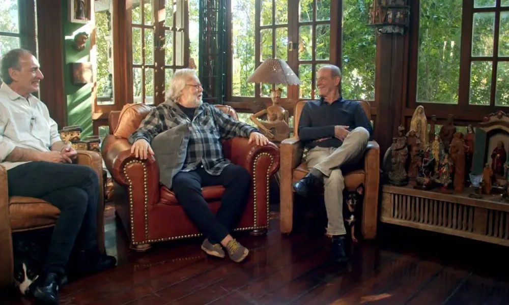 Gérson Conrad, Willy Verdaguer, Ney Matogrosso, cada um em uma poltrona, conversando e rindo durante filmagem do documentário "Primavera nos Dentes: a História dos Secos & Molhados".