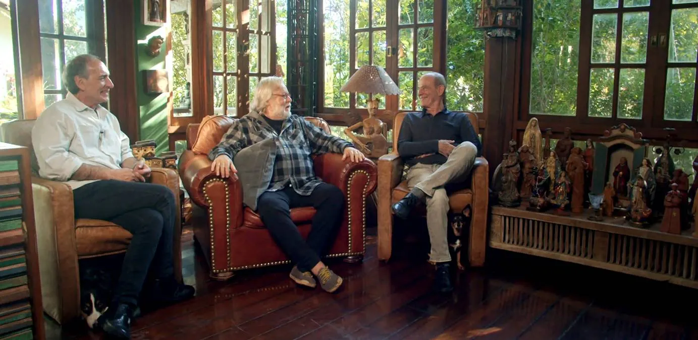 Gérson Conrad, Willy Verdaguer, Ney Matogrosso, cada um em uma poltrona, conversando e rindo durante filmagem do documentário "Primavera nos Dentes: a História dos Secos & Molhados".