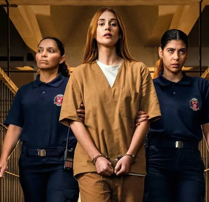 Marina Ruy Barbosa ao centro com roupas de presídio, caracterizada como Suzane von Richtofen, em imagem promocional, cercada por duas carcereiras, para a série "Tremembé", caminhando nos corredores do presídio algemada.