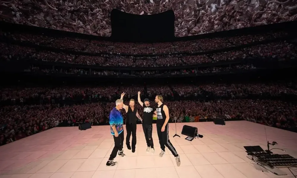 Membros do U2 em show Live at Sphere em 2024, de costas para o palco, tirando foto com o público.