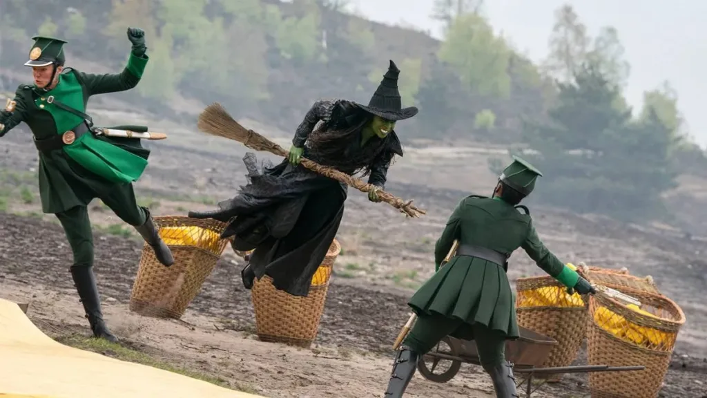 Cynthia Erivo como a bruxa Elphaba, montada em uma vassoura, derrubando guardas na construção da estrada de tijolos amarelos no início da segunda parte de 'Wicked' (2025).
