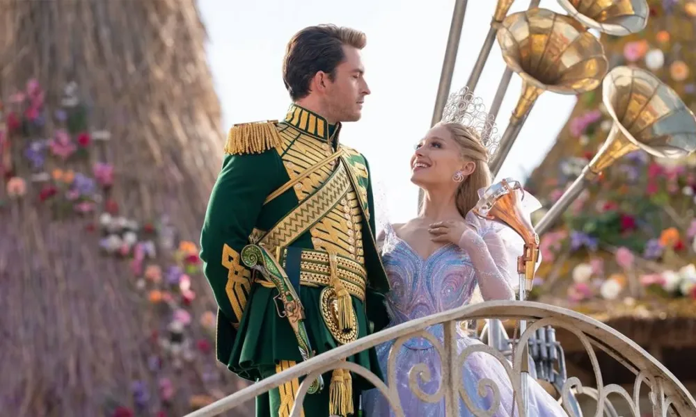 Jonathan Bailey (esquerda) e Ariana Grande (direita) como Fiyero e Glinda na segunda parte de "Wicked" (2025). Eles estão num cortejo em Oz, na frente de um boneco de vime ao fundo. Eles se olham.