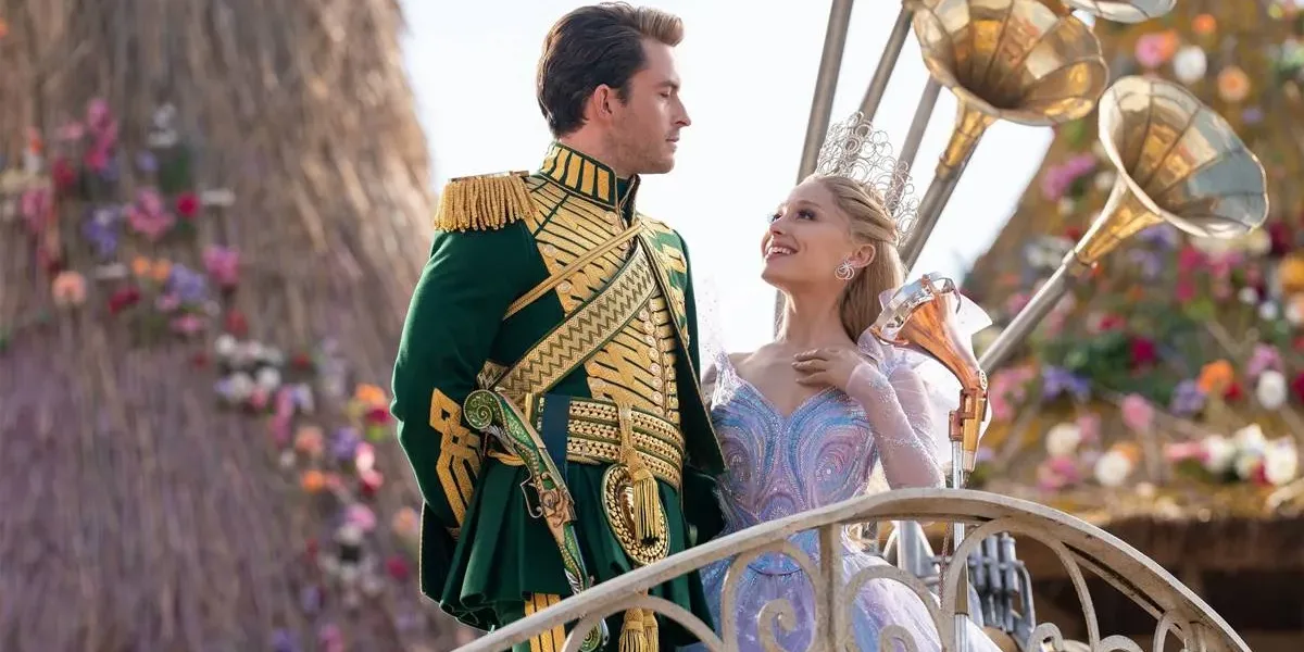 Jonathan Bailey (esquerda) e Ariana Grande (direita) como Fiyero e Glinda na segunda parte de "Wicked" (2025). Eles estão num cortejo em Oz, na frente de um boneco de vime ao fundo. Eles se olham.