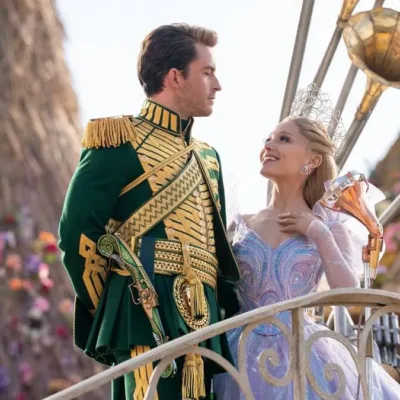Jonathan Bailey (esquerda) e Ariana Grande (direita) como Fiyero e Glinda na segunda parte de "Wicked" (2025). Eles estão num cortejo em Oz, na frente de um boneco de vime ao fundo. Eles se olham.