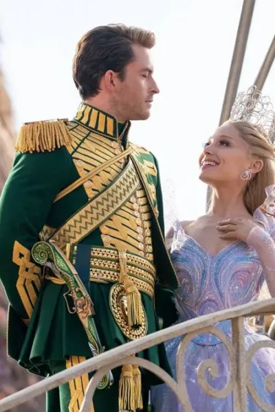 Jonathan Bailey (esquerda) e Ariana Grande (direita) como Fiyero e Glinda na segunda parte de "Wicked" (2025). Eles estão num cortejo em Oz, na frente de um boneco de vime ao fundo. Eles se olham.