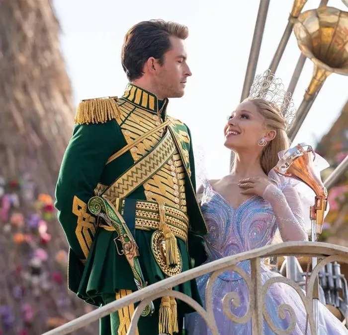 Jonathan Bailey (esquerda) e Ariana Grande (direita) como Fiyero e Glinda na segunda parte de "Wicked" (2025). Eles estão num cortejo em Oz, na frente de um boneco de vime ao fundo. Eles se olham.