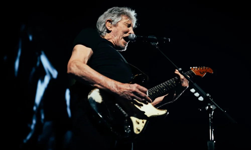 Roger Waters no centro da imagem ao microfone com uma guitarra em apresentação ao vivo