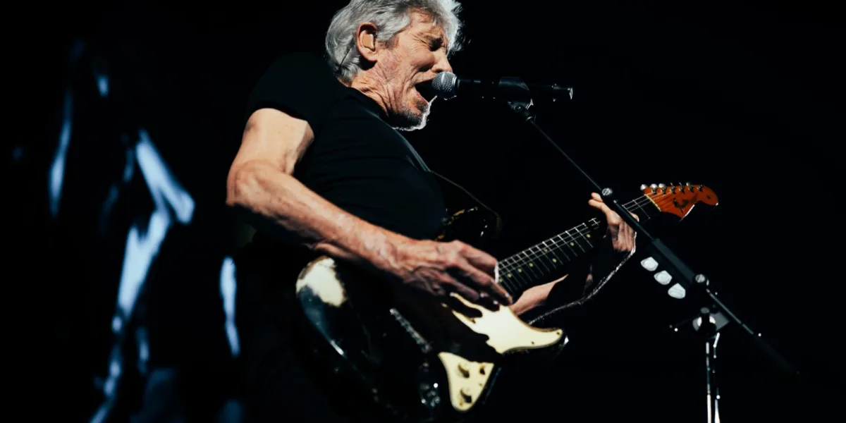 Roger Waters no centro da imagem ao microfone com uma guitarra em apresentação ao vivo