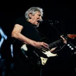 Roger Waters no centro da imagem ao microfone com uma guitarra em apresentação ao vivo