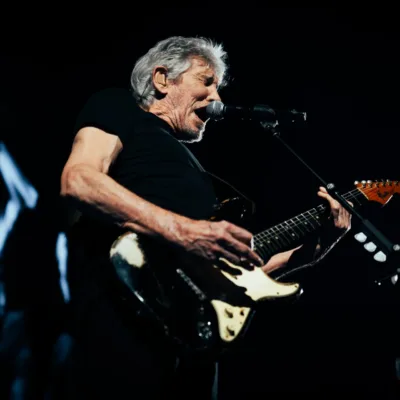 Roger Waters no centro da imagem ao microfone com uma guitarra em apresentação ao vivo