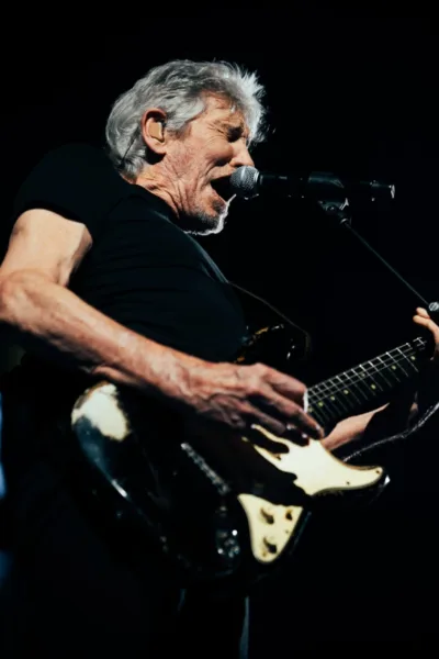 Roger Waters no centro da imagem ao microfone com uma guitarra em apresentação ao vivo