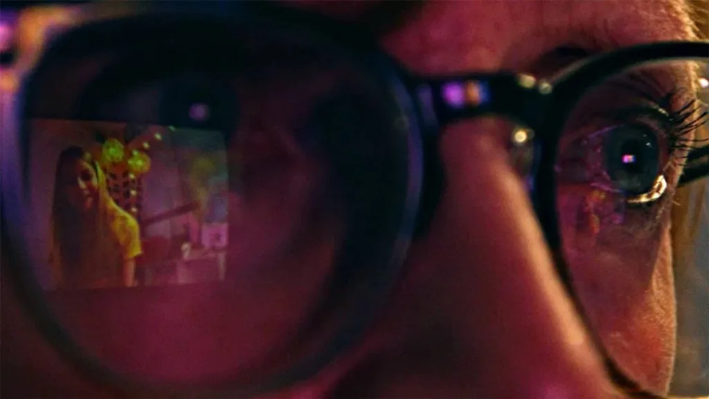 A Garota Artificial | Suspense Eletrizante e Uma Obra Criativa Sobre a IA Que nos Cerca 1 Câmera aproximada no rosto de uma pessoa observando uma tela com um rosto de uma garota gerada por IA, refletida em seus óculos. Imagem do filme "A Garota Artificial".