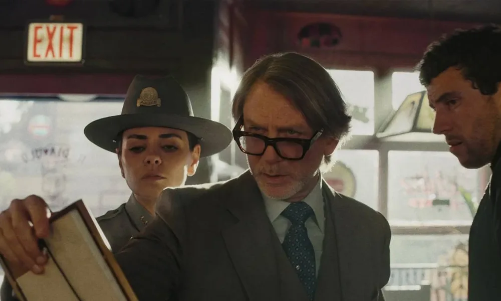 Daniel Craig como o protagonista do terceiro filme da série "Knives Out". Ele segura um papel e olha com seus óculos levemente caídos, um homem vê atrás dele também e também a delegada.