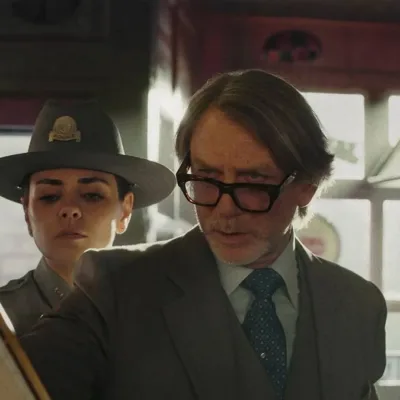 Daniel Craig como o protagonista do terceiro filme da série "Knives Out". Ele segura um papel e olha com seus óculos levemente caídos, um homem vê atrás dele também e também a delegada.