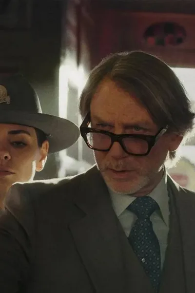 Daniel Craig como o protagonista do terceiro filme da série "Knives Out". Ele segura um papel e olha com seus óculos levemente caídos, um homem vê atrás dele também e também a delegada.