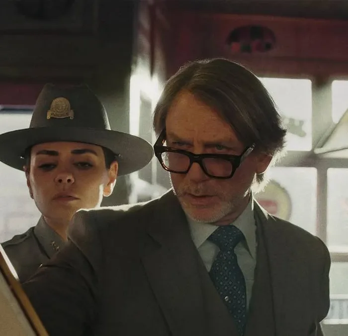 Daniel Craig como o protagonista do terceiro filme da série "Knives Out". Ele segura um papel e olha com seus óculos levemente caídos, um homem vê atrás dele também e também a delegada.