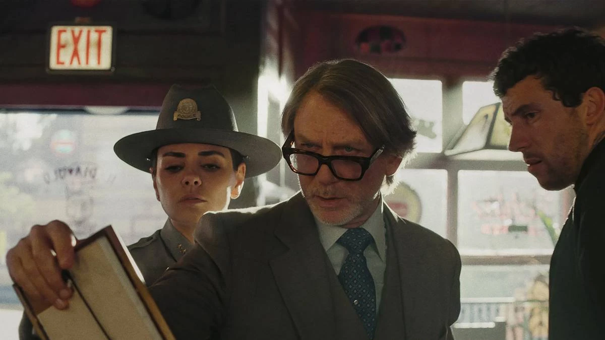 Daniel Craig como o protagonista do terceiro filme da série "Knives Out". Ele segura um papel e olha com seus óculos levemente caídos, um homem vê atrás dele também e também a delegada.