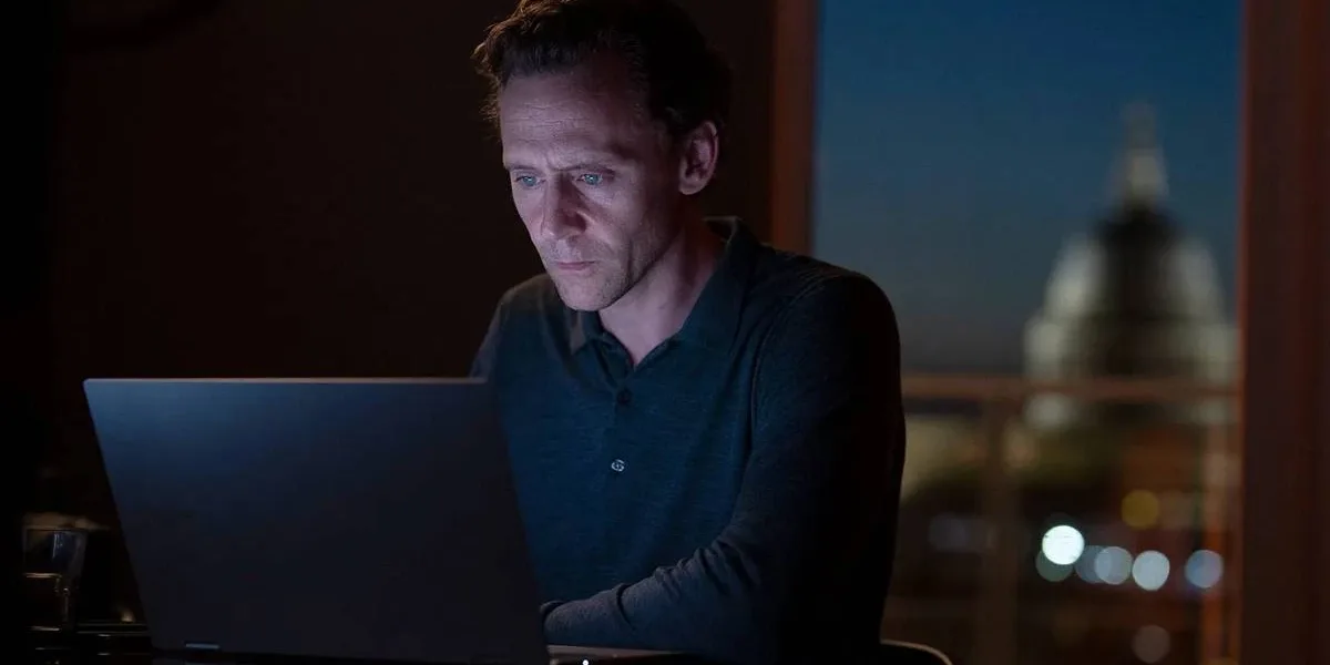 Tom Hiddleston como o protagonista de "O Gerente Noturno" no primeiro episódio da segunda temporada da série. Ele está mexendo em um computador, que é a única iluminação do ambiente junto com a janela ao fundo, no início da noite, em um ambiente urbano.