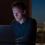 Tom Hiddleston como o protagonista de "O Gerente Noturno" no primeiro episódio da segunda temporada da série. Ele está mexendo em um computador, que é a única iluminação do ambiente junto com a janela ao fundo, no início da noite, em um ambiente urbano.