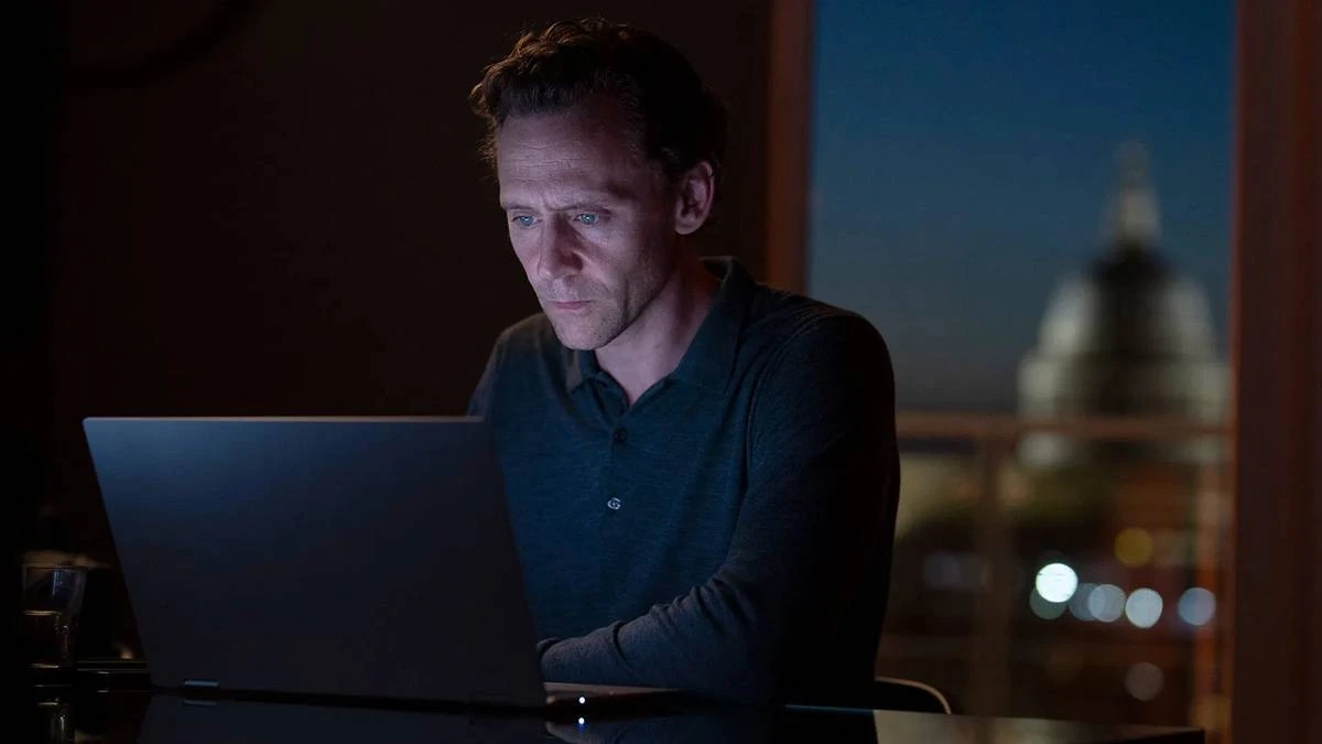 Tom Hiddleston como o protagonista de "O Gerente Noturno" no primeiro episódio da segunda temporada da série. Ele está mexendo em um computador, que é a única iluminação do ambiente junto com a janela ao fundo, no início da noite, em um ambiente urbano.