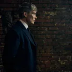 Cillian Murphy caracterizado como Thomas Shelby para o filme de fechamento da franquia "Peaky Blinders", ambientado na 2ª Guerra Mundial, "O Homem Imortal". Ele está de perfil, mãos para trás, com um muro de tijolos ao fundo.
