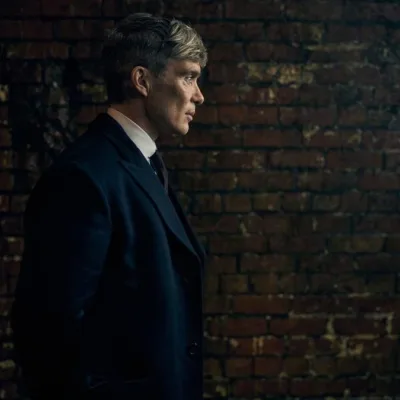 Cillian Murphy caracterizado como Thomas Shelby para o filme de fechamento da franquia "Peaky Blinders", ambientado na 2ª Guerra Mundial, "O Homem Imortal". Ele está de perfil, mãos para trás, com um muro de tijolos ao fundo.