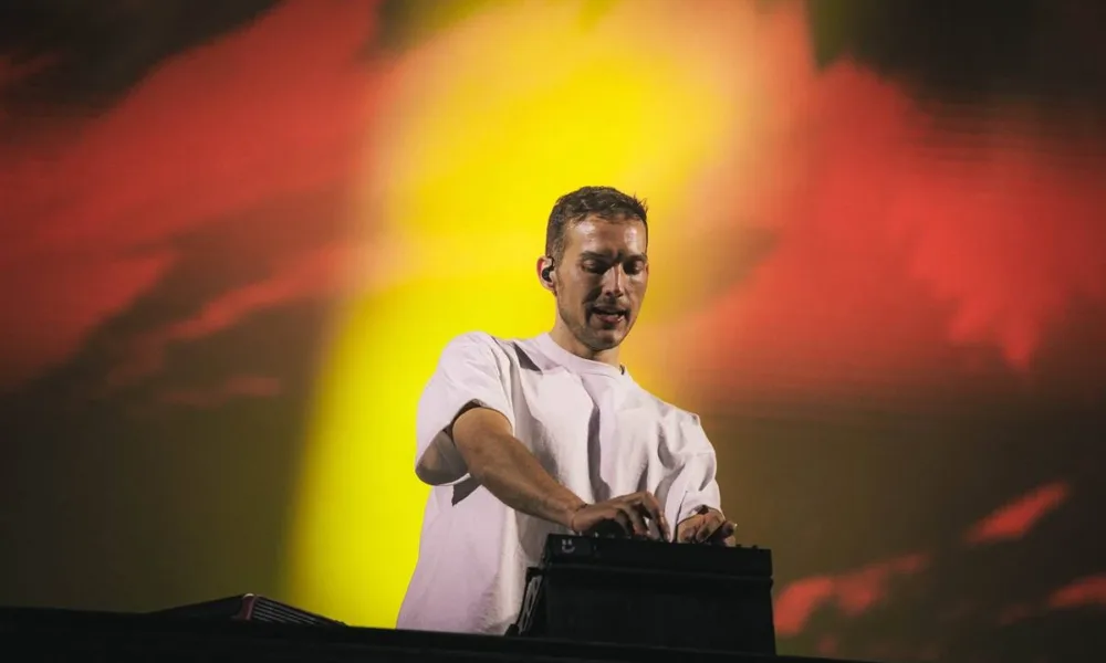 Kygo em foto retirada de um ângulo abaixo do palco, o DJ em seu set em fundo vermelho e amarelo luzes quase opacas, dando destaque ao artista.