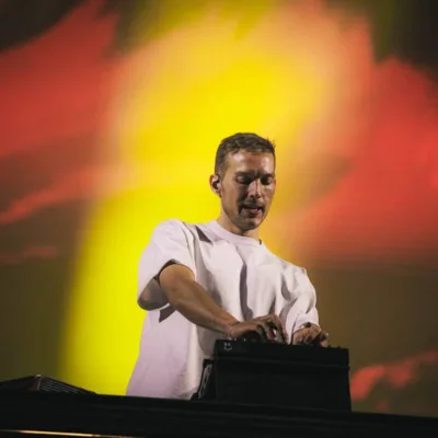 Kygo em foto retirada de um ângulo abaixo do palco, o DJ em seu set em fundo vermelho e amarelo luzes quase opacas, dando destaque ao artista.