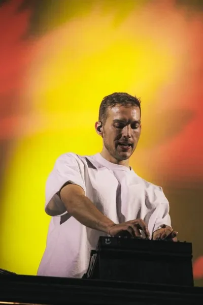 Kygo em foto retirada de um ângulo abaixo do palco, o DJ em seu set em fundo vermelho e amarelo luzes quase opacas, dando destaque ao artista.