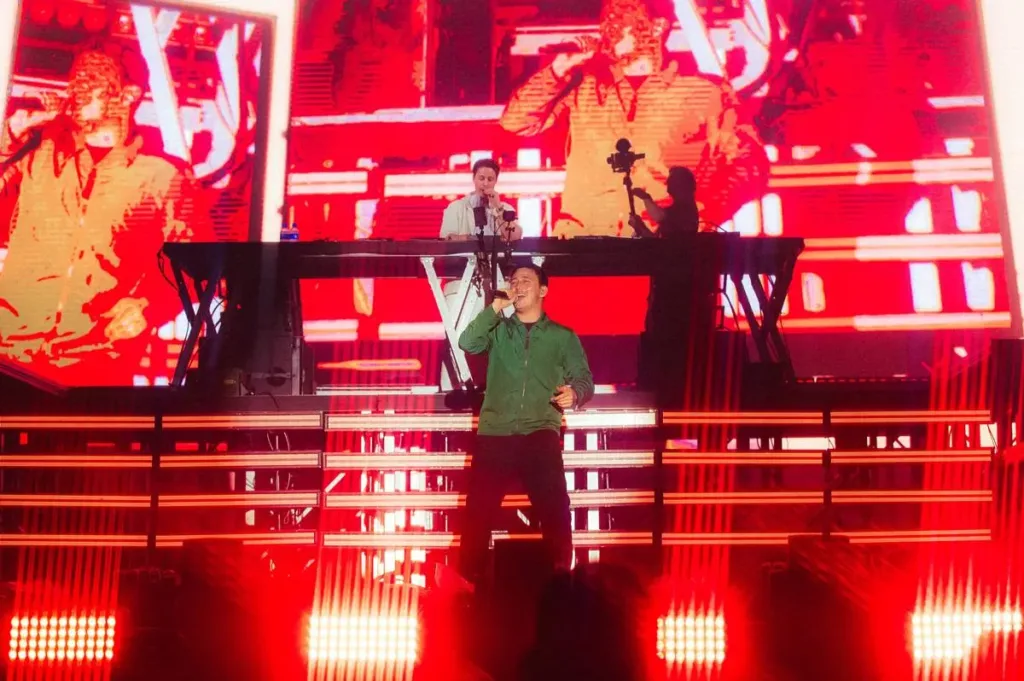 Kygo no palco Perry's no Lollapalooza 2026, microfone na mão, de pé, centralizado em foto com luzes vermelhas ao fundo dando profundidade à apresentação.