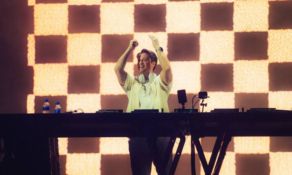 Kygo no palco Perry's no Lollapalooza 2026, microfone na mão, de pé, centralizado em foto atrás de seu aparelho de mixagem, mãos levantadas batendo palmas e com um sorriso no rosto.