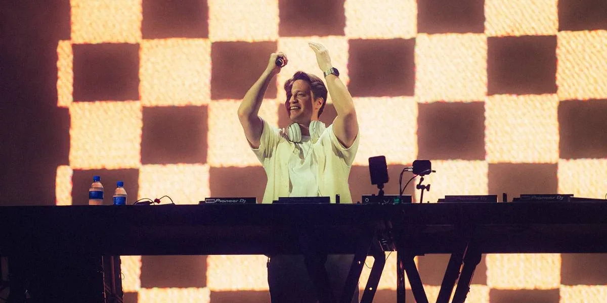 Kygo no palco Perry's no Lollapalooza 2026, microfone na mão, de pé, centralizado em foto atrás de seu aparelho de mixagem, mãos levantadas batendo palmas e com um sorriso no rosto.