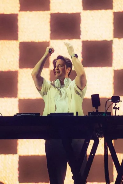 Kygo no palco Perry's no Lollapalooza 2026, microfone na mão, de pé, centralizado em foto atrás de seu aparelho de mixagem, mãos levantadas batendo palmas e com um sorriso no rosto.