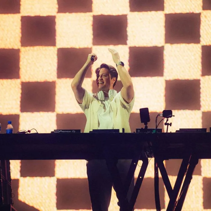 Kygo no palco Perry's no Lollapalooza 2026, microfone na mão, de pé, centralizado em foto atrás de seu aparelho de mixagem, mãos levantadas batendo palmas e com um sorriso no rosto.