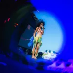 Captura de tela através de lente azul focando em Agnes Nunes durante seu show no Lollapalooza 2026. Cantoar está usando um vestido com top e saia verde.