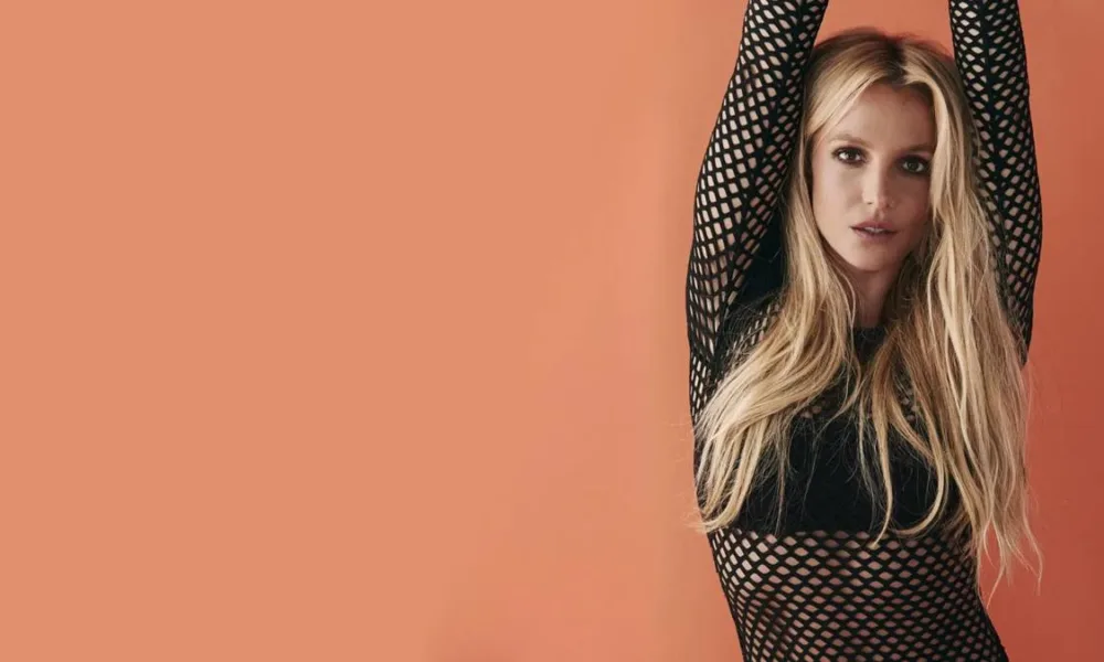 Britney Spears à direita da tela com braços erguidos em dança, encarando a câmera, em parede de fundo salmão em ensaio para o documentário "Britney: No Filter".