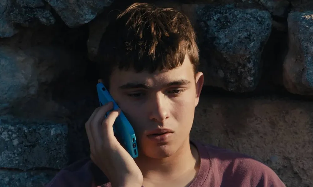 Eloy Pohu como Enzo no filme autointitulado, segurando um celular e preocupado sentado contra um muro de pedra.
