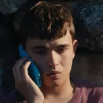 Eloy Pohu como Enzo no filme autointitulado, segurando um celular e preocupado sentado contra um muro de pedra.