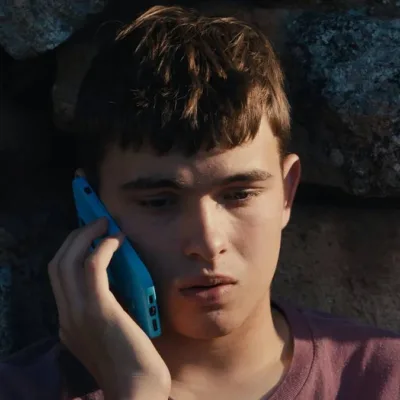 Eloy Pohu como Enzo no filme autointitulado, segurando um celular e preocupado sentado contra um muro de pedra.