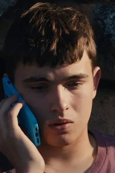 Eloy Pohu como Enzo no filme autointitulado, segurando um celular e preocupado sentado contra um muro de pedra.